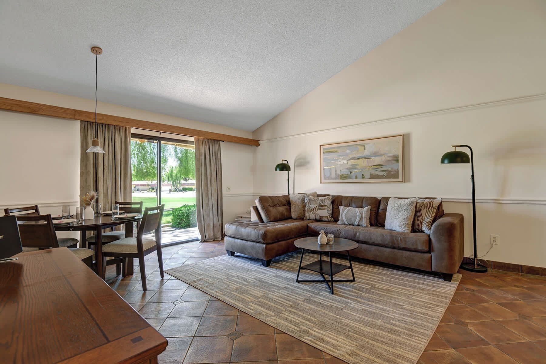 A living room with a brown leather sofa, a dining table with chairs, a coffee table, and a large window with curtains.