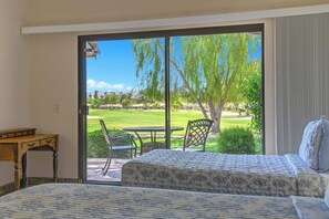 A hotel room with a bed, a small table, and a chair, with a view of a golf course through a sliding glass door. - Relaxing 2 Bedroom Palm Resort Escape (Palm Desert)