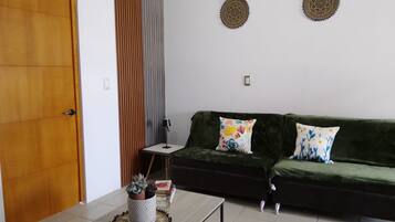 Un salon avec un canapé vert foncé, deux coussins à motifs, une petite table avec une lampe et un élément décoratif mural.