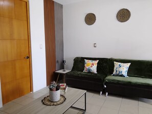A living room with a dark green sofa, two patterned cushions, a small table with a lamp, and a decorative wall piece.