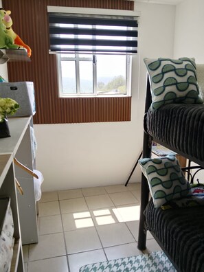 A room with a bunk bed, a window with blinds, a radiator, and a shelf with various items.