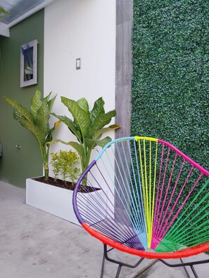 A colorful chair in a planter box, a green wall with foliage, and a white wall with a picture frame.