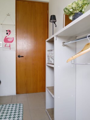 Una habitación con una puerta de madera, un estante blanco con una cuchara de madera colgante, una lámpara negra y una planta fijada a la pared.