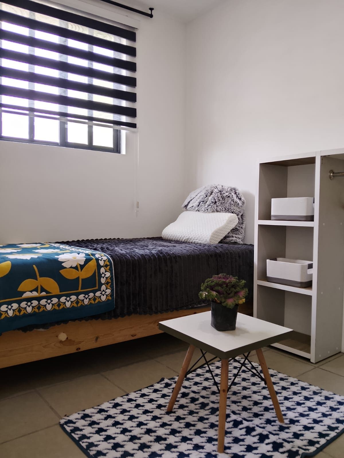 Ein Schlafzimmer mit einem Bett, einem Nachttisch mit Pflanze, einem Fenster mit Jalousien und einem Kleiderschrank.