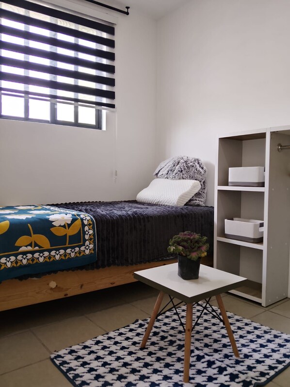 A bedroom with a bed, a bedside table with a plant, a window with blinds, and a wardrobe.