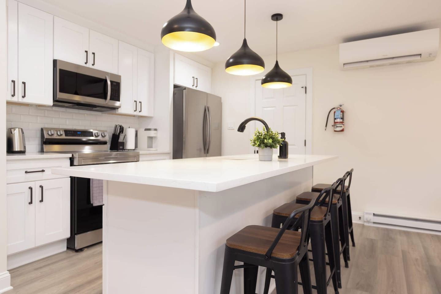 Una cocina moderna con armarios blancos, electrodomésticos de acero inoxidable, una isla central con taburetes de bar, iluminación colgante y una unidad de aire acondicionado a la vista.