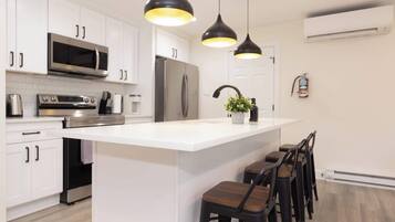Una cocina moderna con armarios blancos, electrodomésticos de acero inoxidable, una isla central con taburetes de bar, iluminación colgante y una unidad de aire acondicionado a la vista.