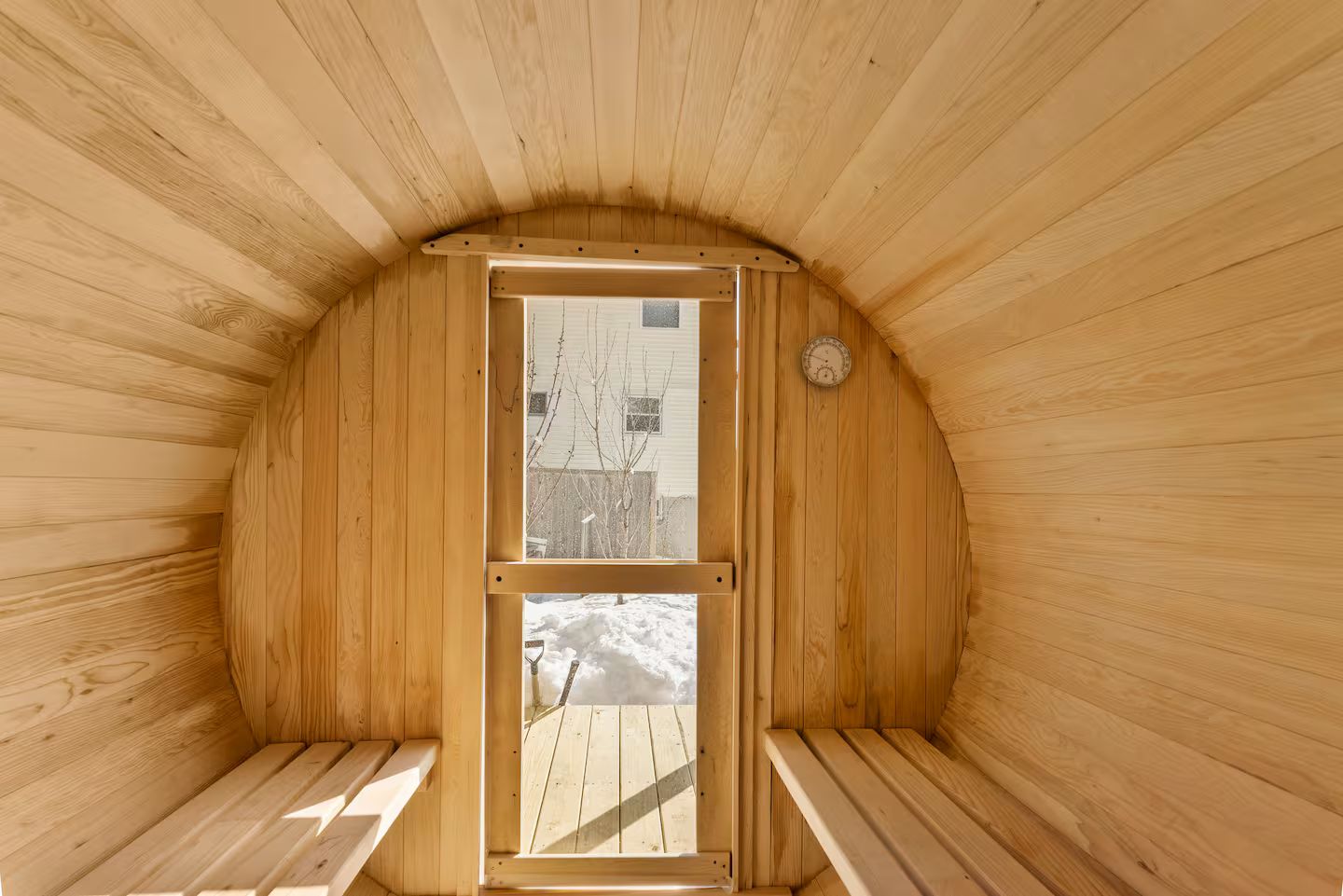 A sauna with wooden walls and benches, a door with a view of the outside, and a wall-mounted thermometer.