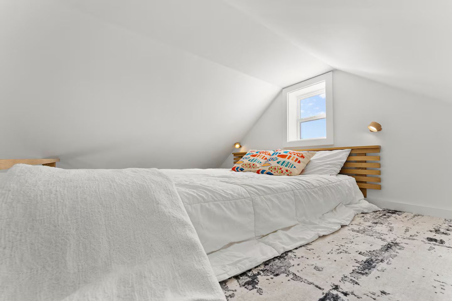 A bedroom with a sloped ceiling, a bed with white bedding, a colorful pillow, and a small window.