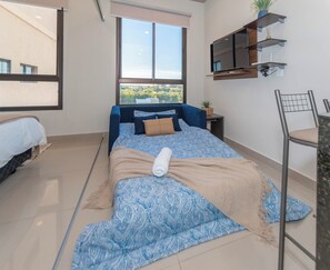 A bedroom with a bed, a window with a city view, a TV mounted on the wall, and a chair. - Zuba IV Studio | Rooftop Pool & WiFi (Luque)