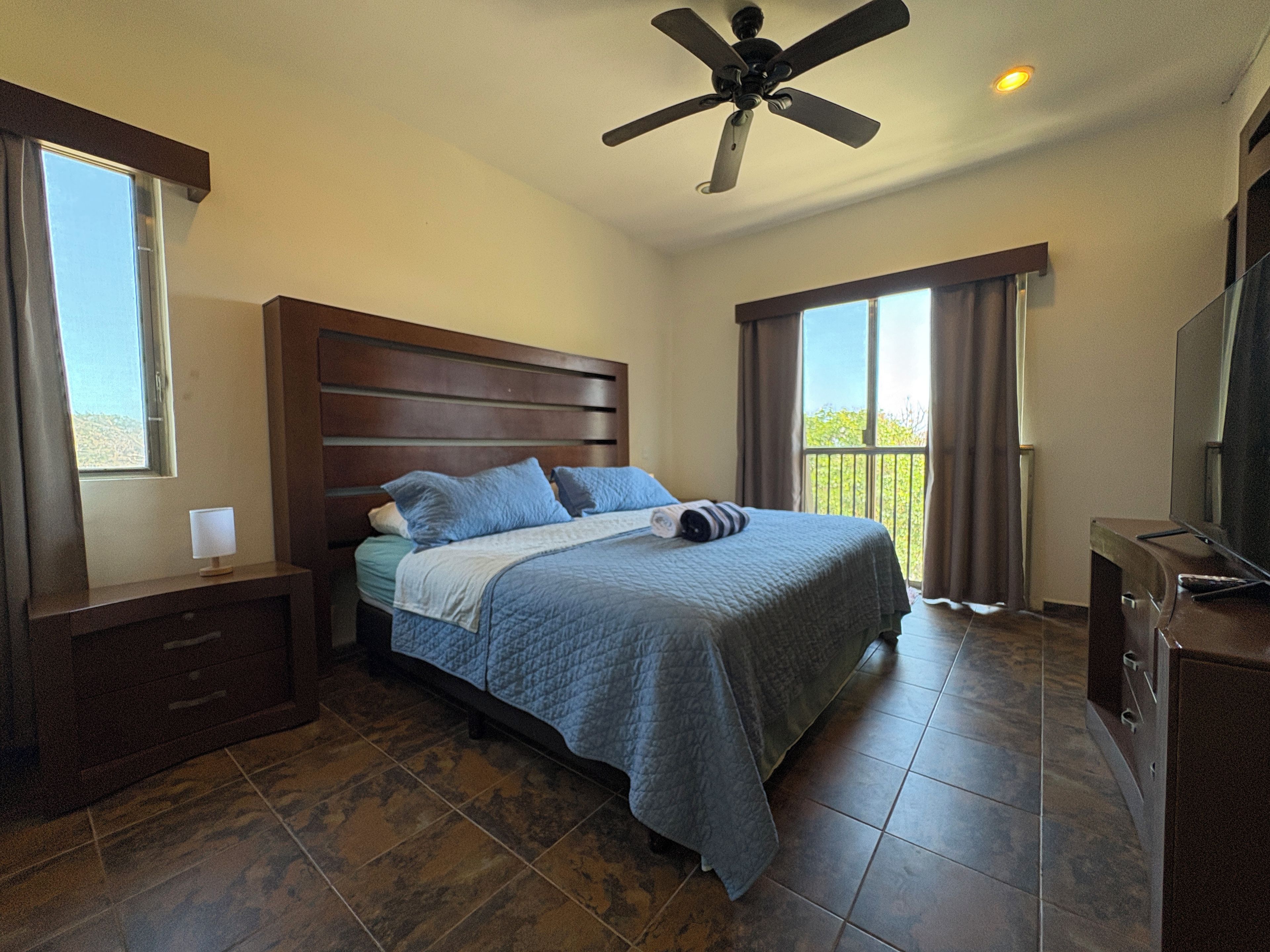 A hotel room with a bed, a ceiling fan, a TV, and a balcony.