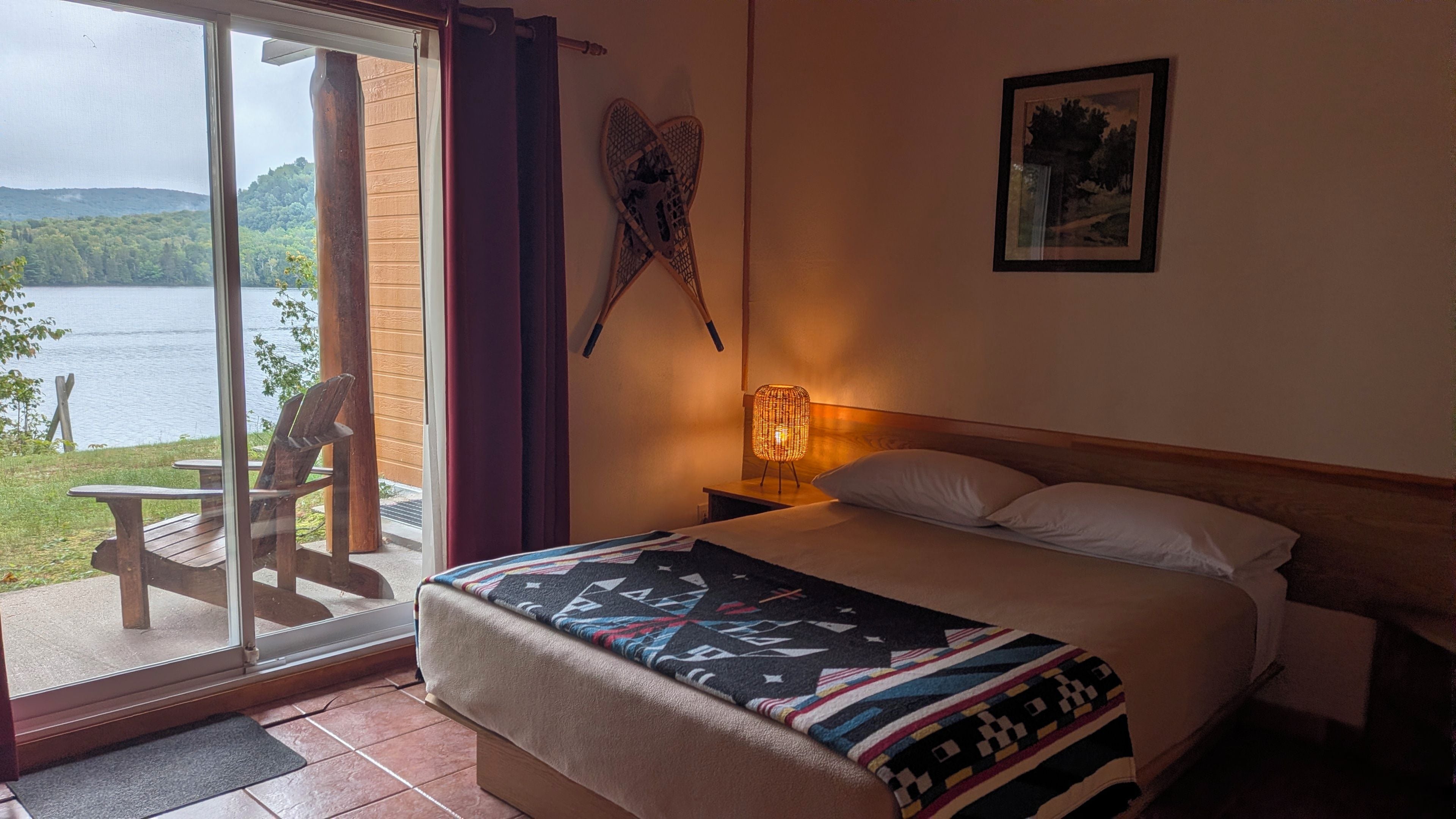 Une chambre d’hôtel avec un lit, une table de chevet, une lampe, un tableau au mur et une vue sur un lac, visible à travers une porte vitrée.