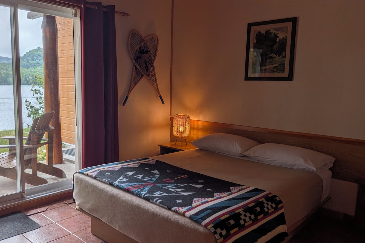 A hotel room with a bed, a nightstand, a lamp, a picture on the wall, and a view of a lake through a glass door.