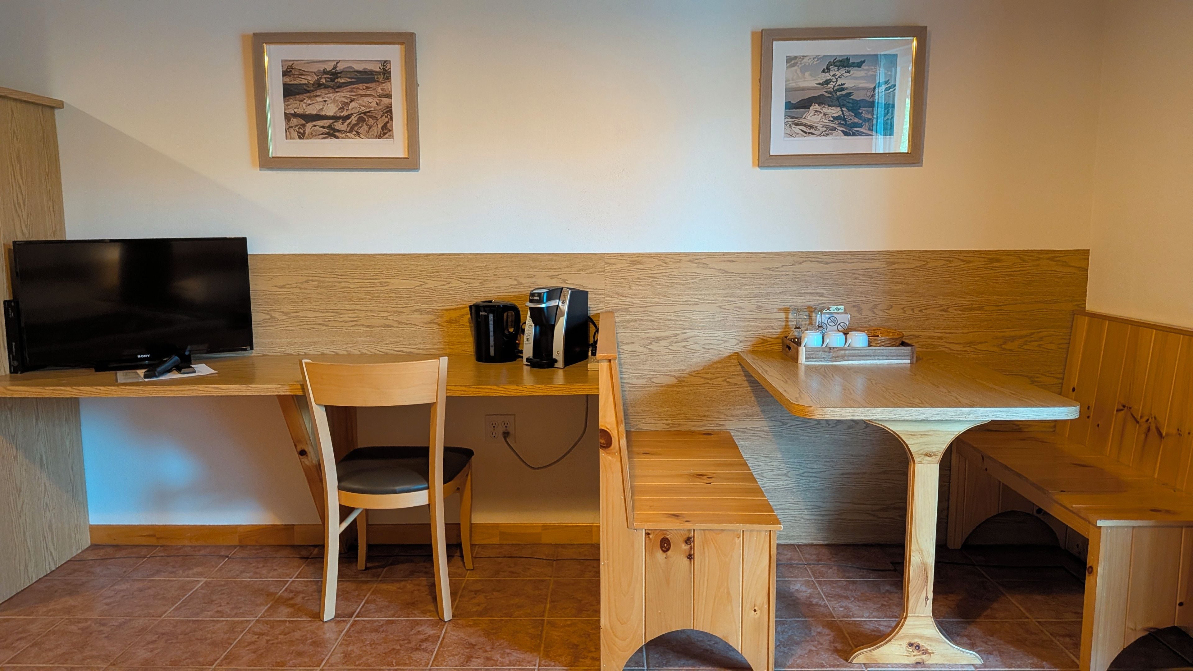 A room with a wooden desk, a chair, a television, a coffee maker, and a bench.
