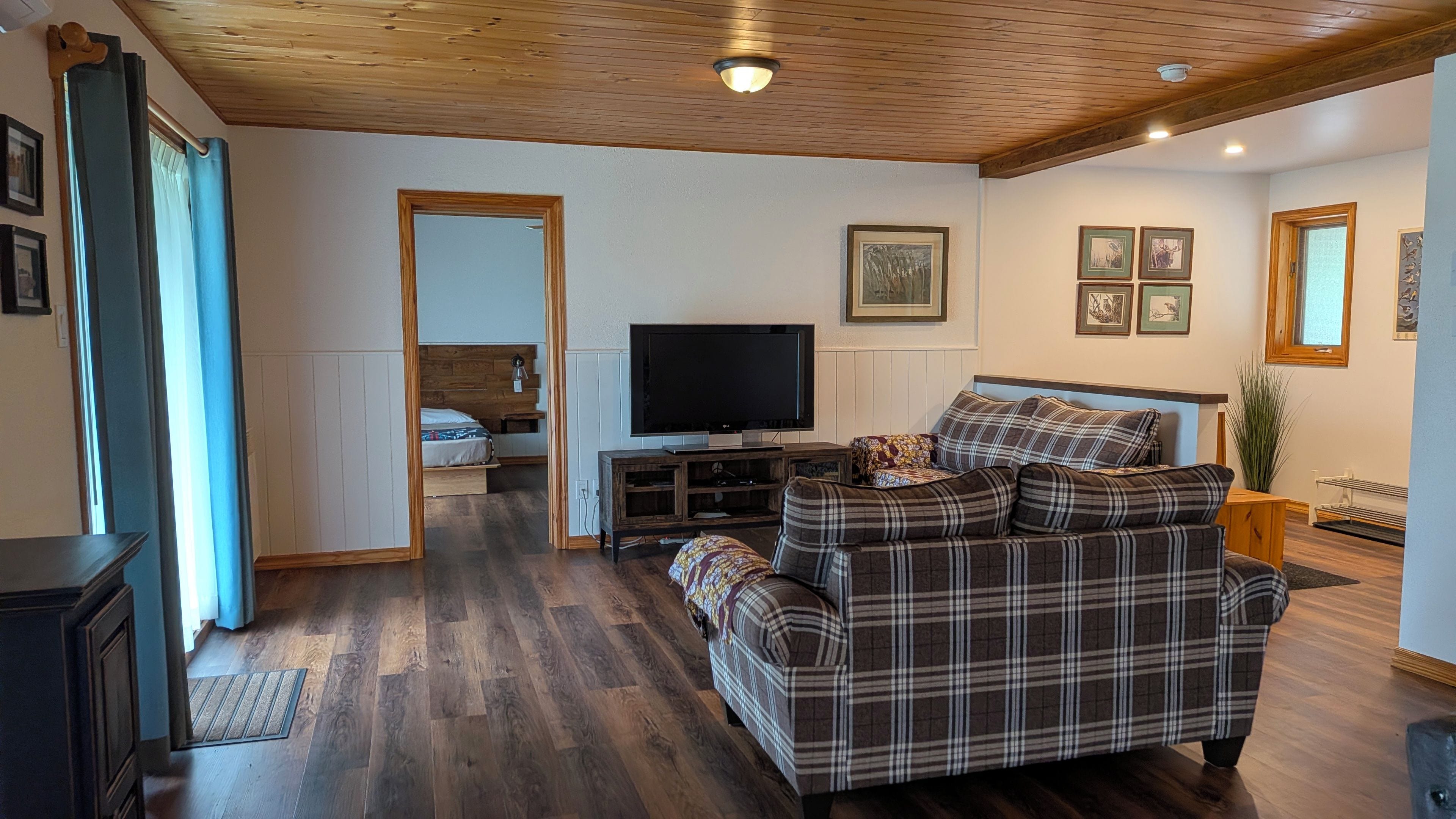 A living room with a wooden ceiling, a television on a stand, a sofa, and a bed in the background.