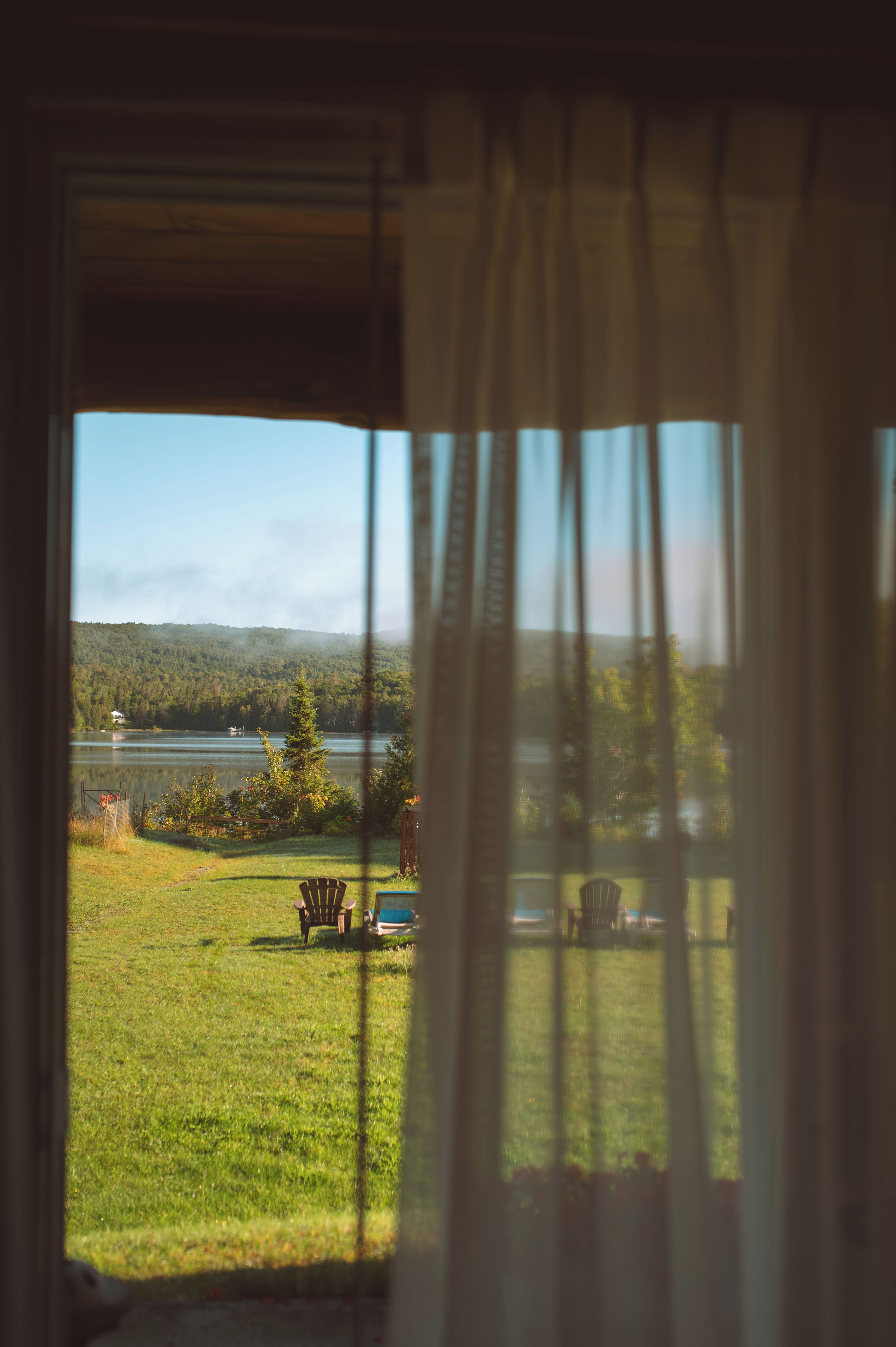 A view through a window with curtains, featuring a grassy lawn, a wooden chair, and a body of water with trees in the background.