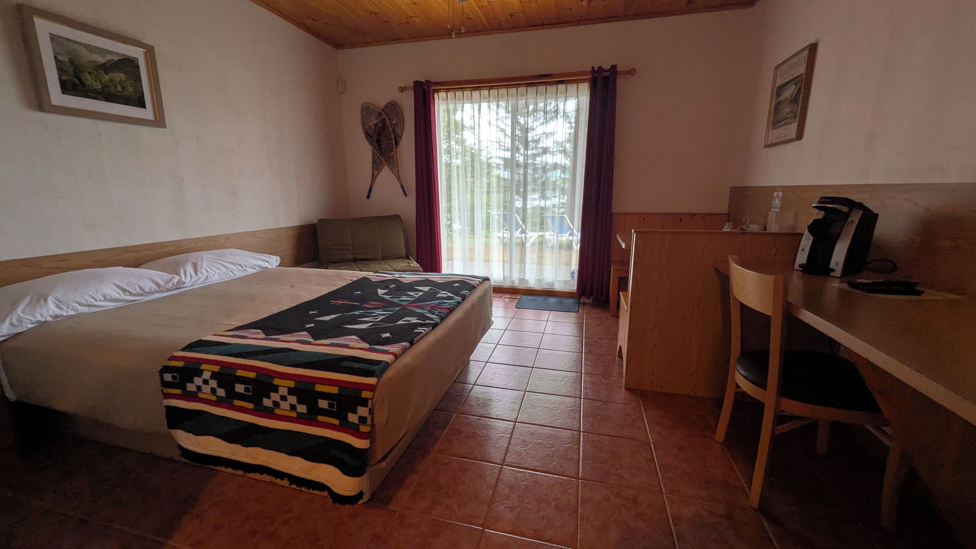 A bedroom with a bed, a desk, and a chair. There is a window with curtains, a door, and a wall-mounted picture.