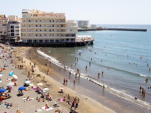 Ein Strand mit einem Steg, Menschen beim Schwimmen und Gebäuden im Hintergrund.