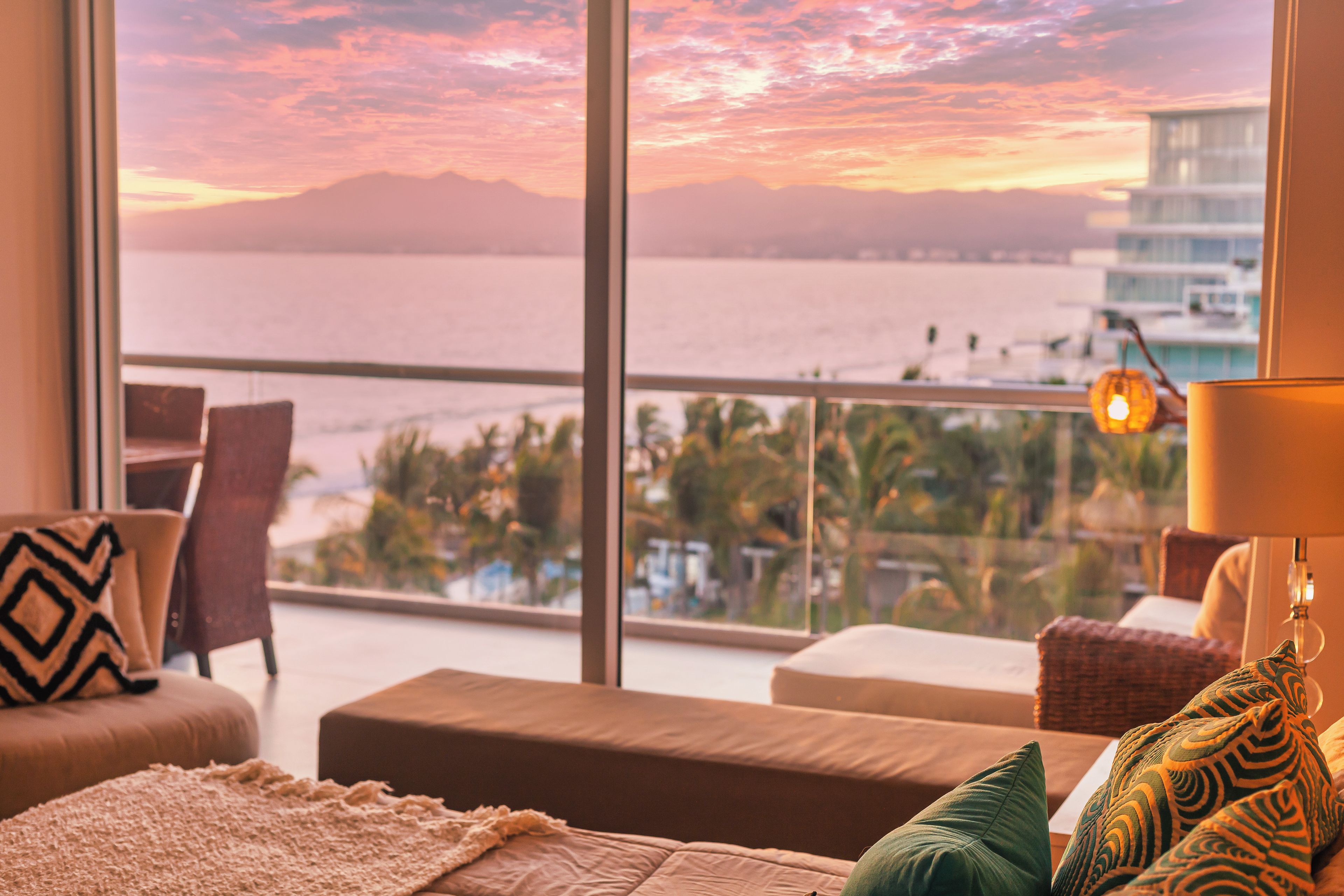 A hotel room with a large window offering a view of the sea and mountains. There is a sofa, a chair, and a lamp on a side table.