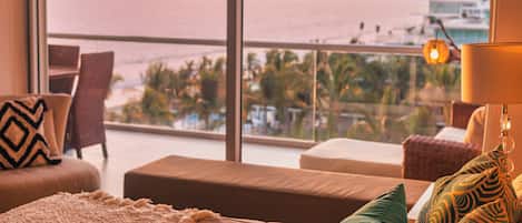 A hotel room with a large window offering a view of the sea and mountains. There is a sofa, a chair, and a lamp on a side table.