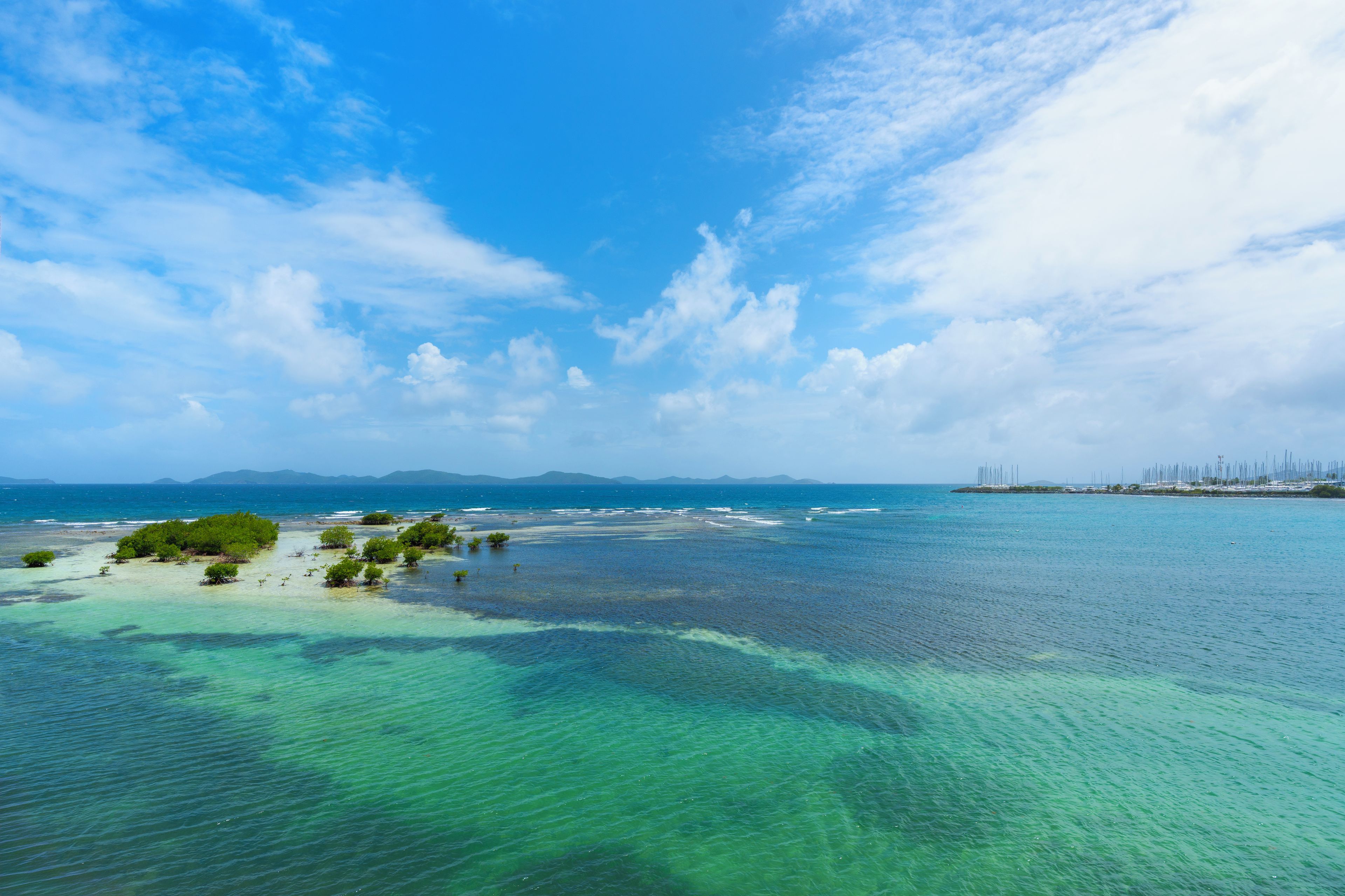 Un paysage côtier avec une eau turquoise et limpide, une petite île couverte d’une végétation luxuriante et, au loin, des reliefs sous un ciel partiellement nuageux.