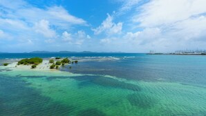 Un paisaje costero con aguas turquesas cristalinas, una pequeña isla con vegetación densa y formaciones terrestres distantes bajo un cielo parcialmente nublado.