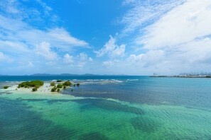 Un paysage côtier avec une eau turquoise et limpide, une petite île couverte d’une végétation luxuriante et, au loin, des reliefs sous un ciel partiellement nuageux.