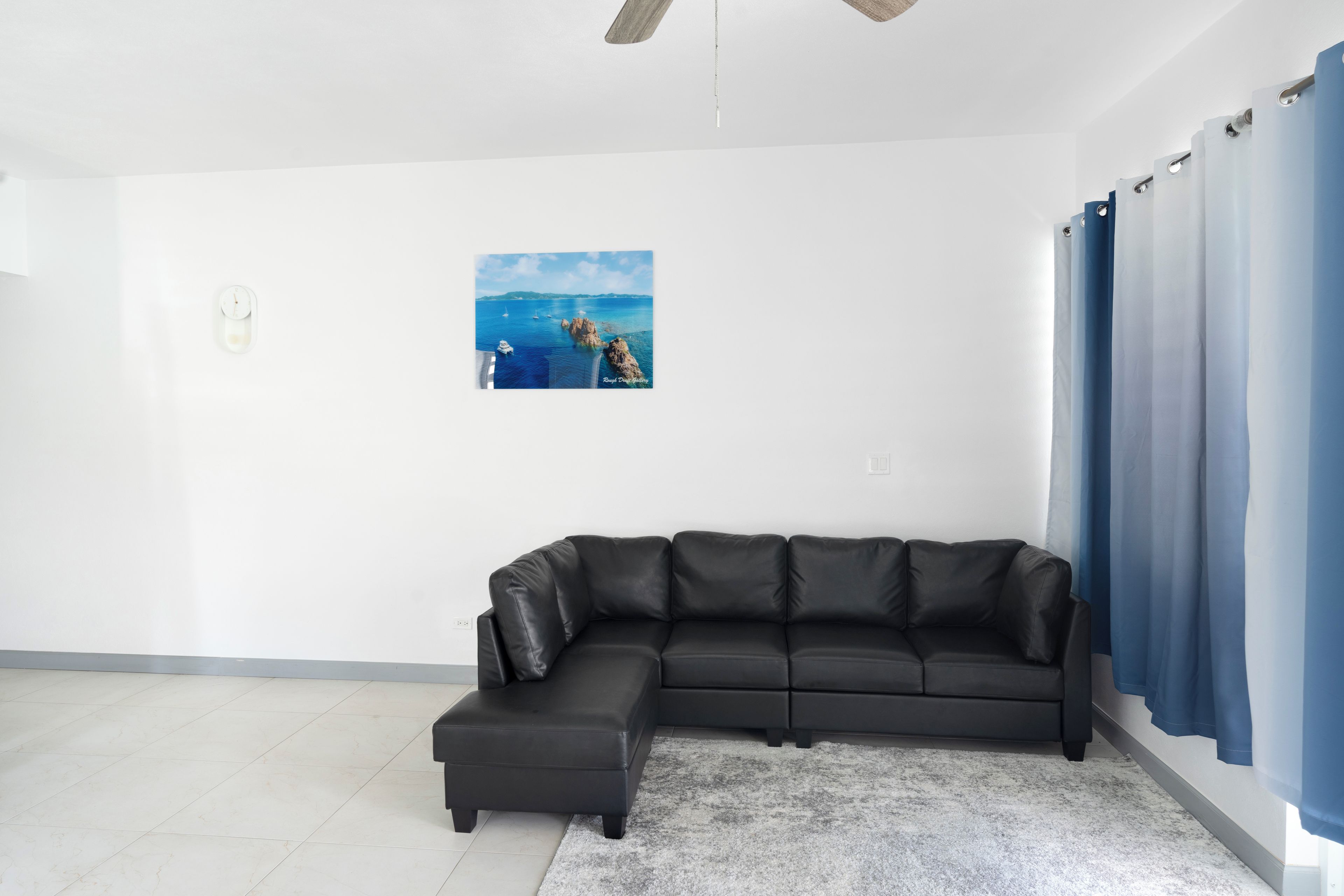 A modern living room with a sectional sofa, a framed coastal landscape painting, and a large window with blue curtains.