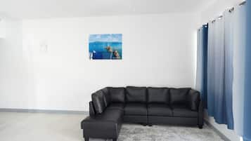 A modern living room with a sectional sofa, a framed coastal landscape painting, and a large window with blue curtains.