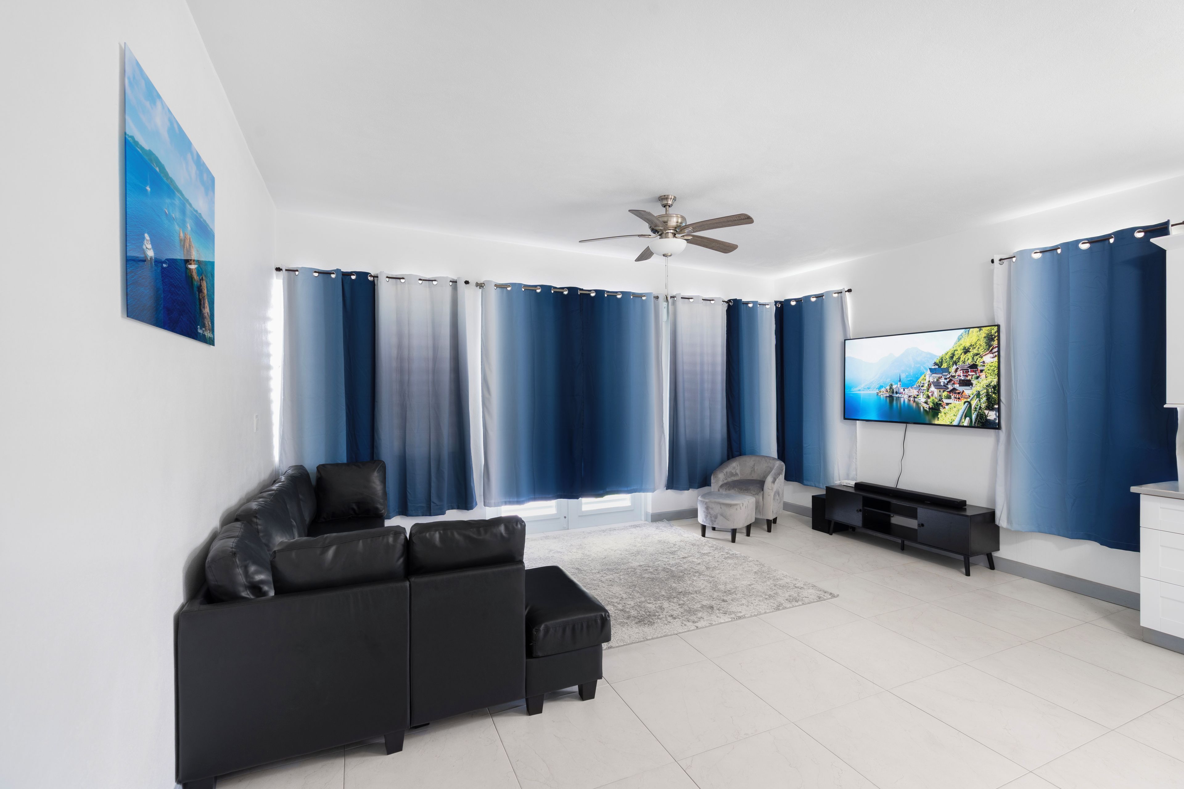 A modern living room with a black sofa, a grey armchair, a flat-screen TV, and a ceiling fan.