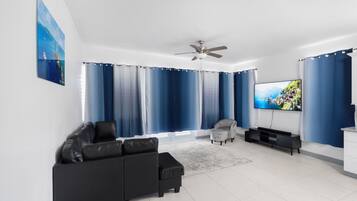 A modern living room with a black sofa, a grey armchair, a flat-screen TV, and a ceiling fan.