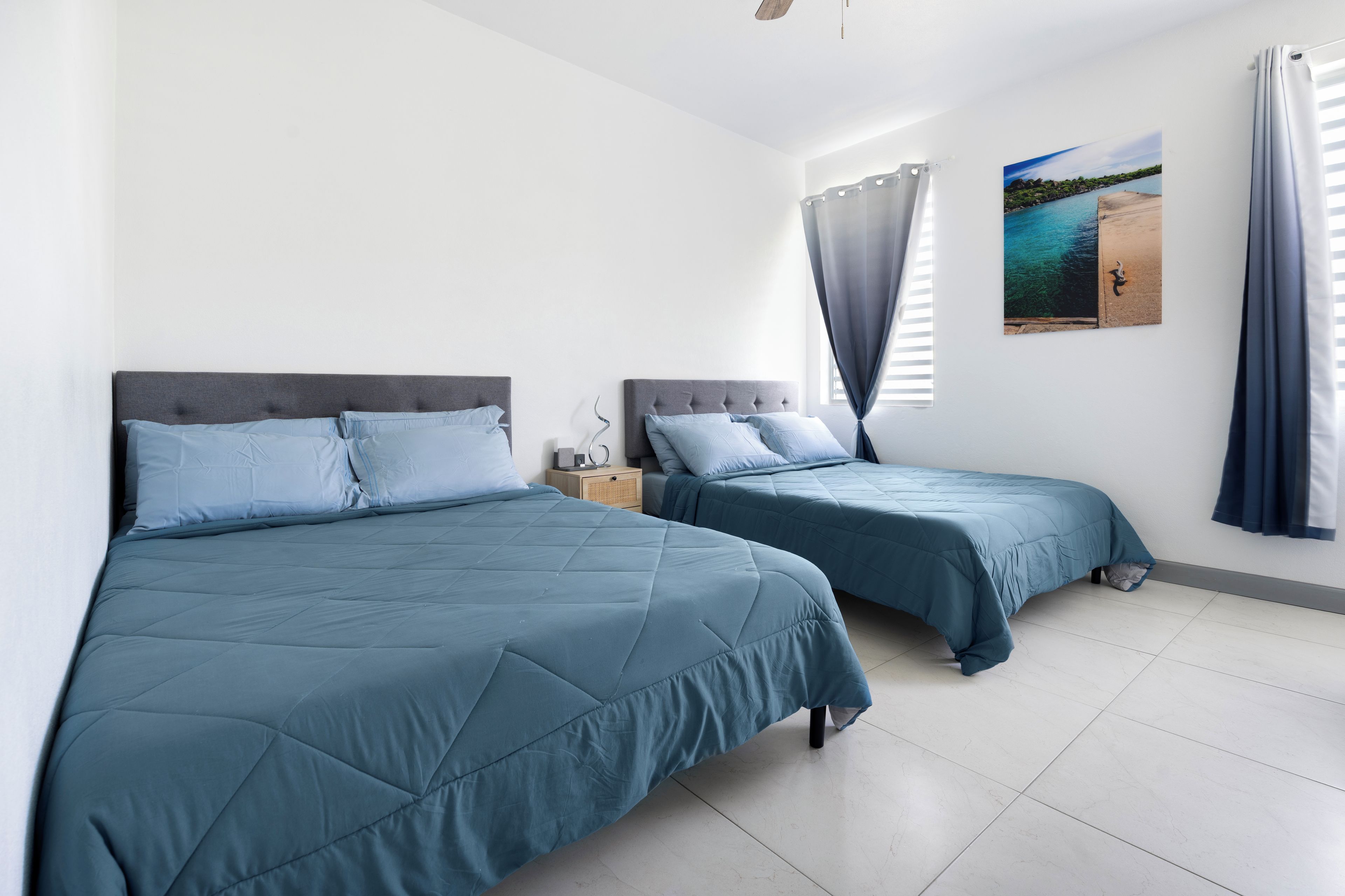 A bedroom with two beds, a nightstand, a window with curtains, and a wall-mounted picture.