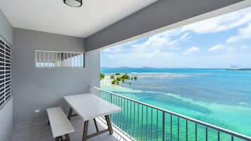 A balcony with a white table and bench, overlooking a clear blue sea and distant land.