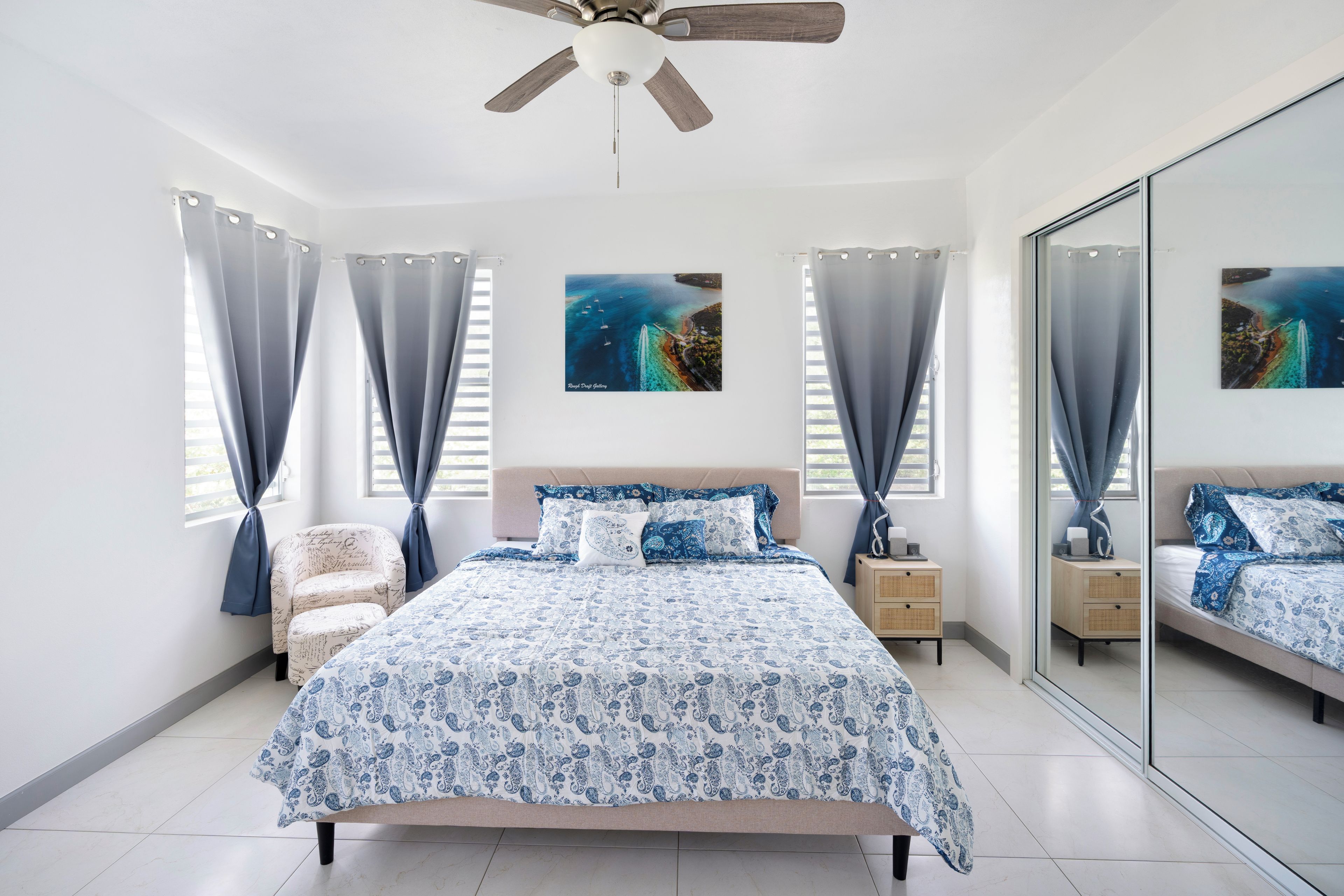 Une chambre à coucher comprenant un lit, des tables de chevet, une chaise, un ventilateur de plafond et deux tableaux accrochés au mur.