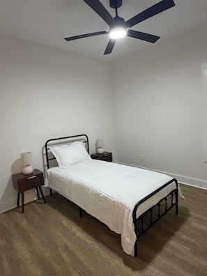 Une chambre avec un lit simple, une table de chevet, une lampe et un ventilateur de plafond.