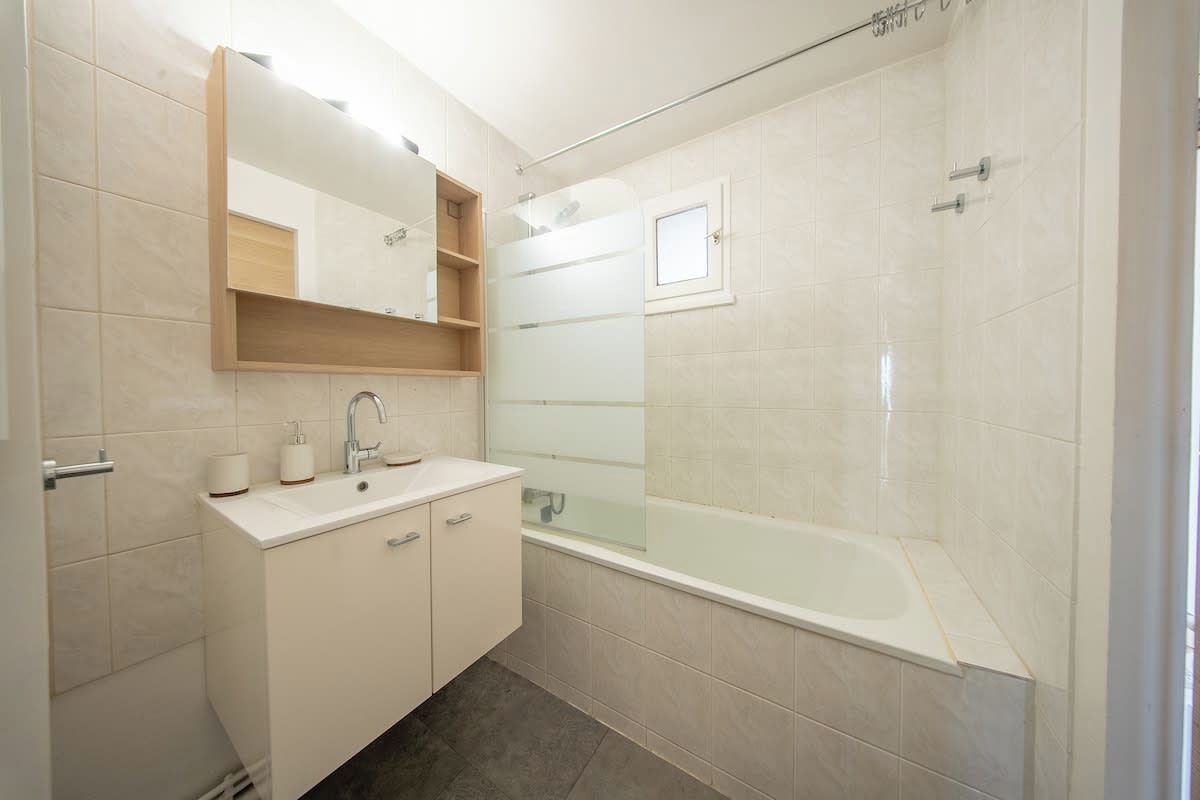 A modern bathroom with a glass-enclosed shower, a white sink with a mirror above, and a beige tiled floor.