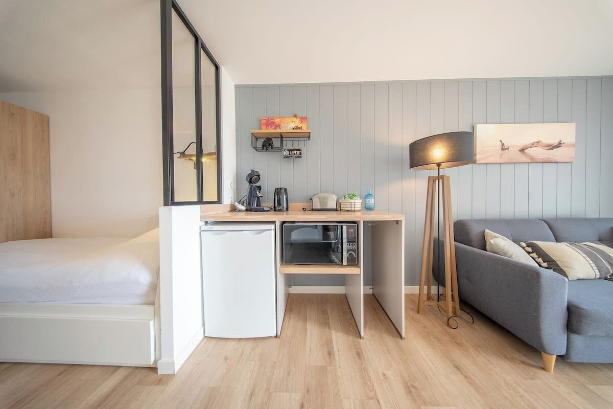 A compact room with a bed, a kitchenette, a desk, a sofa, and a floor lamp.