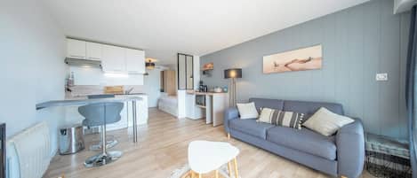 A modern living space with a grey sofa, a dining table, and a kitchen area.