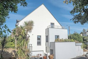 Une maison moderne à deux étages, avec une façade blanche, un balcon à l’étage supérieur et un petit jardin agrémenté de palmiers.
