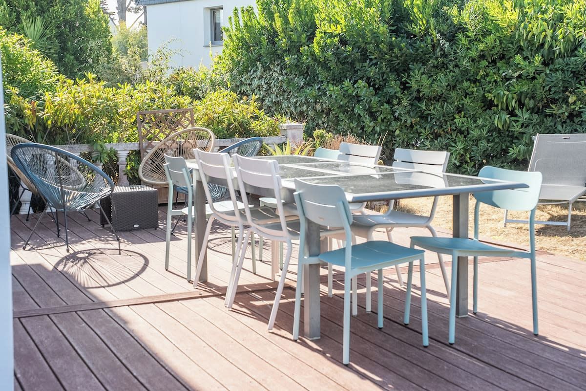 Une terrasse extérieure avec une table et des chaises à plateau en verre, entourée de verdure et avec un bâtiment blanc en arrière-plan.