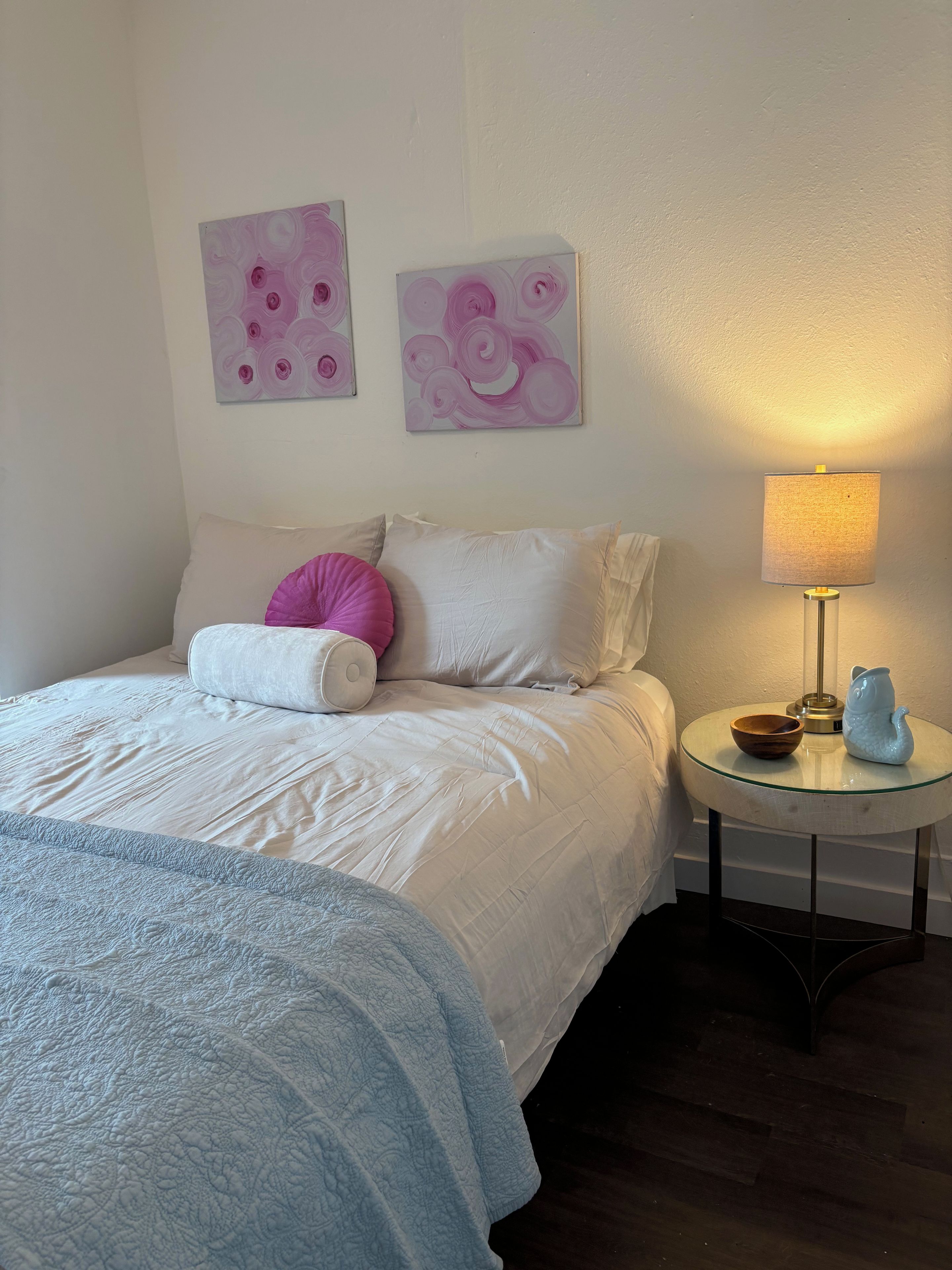Un dormitorio con cama, almohadas, una mesita de noche con lámpara y dos obras de arte en la pared.