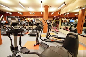 A gym with various exercise machines, including treadmills and elliptical trainers, arranged in a spacious layout with orange flooring and mirrored walls.