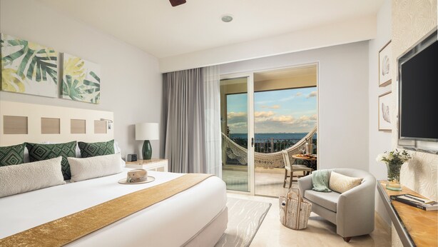 Een moderne hotelkamer met een groot bed, een bank, een bureau en uitzicht op de zee door een glazen deur.
