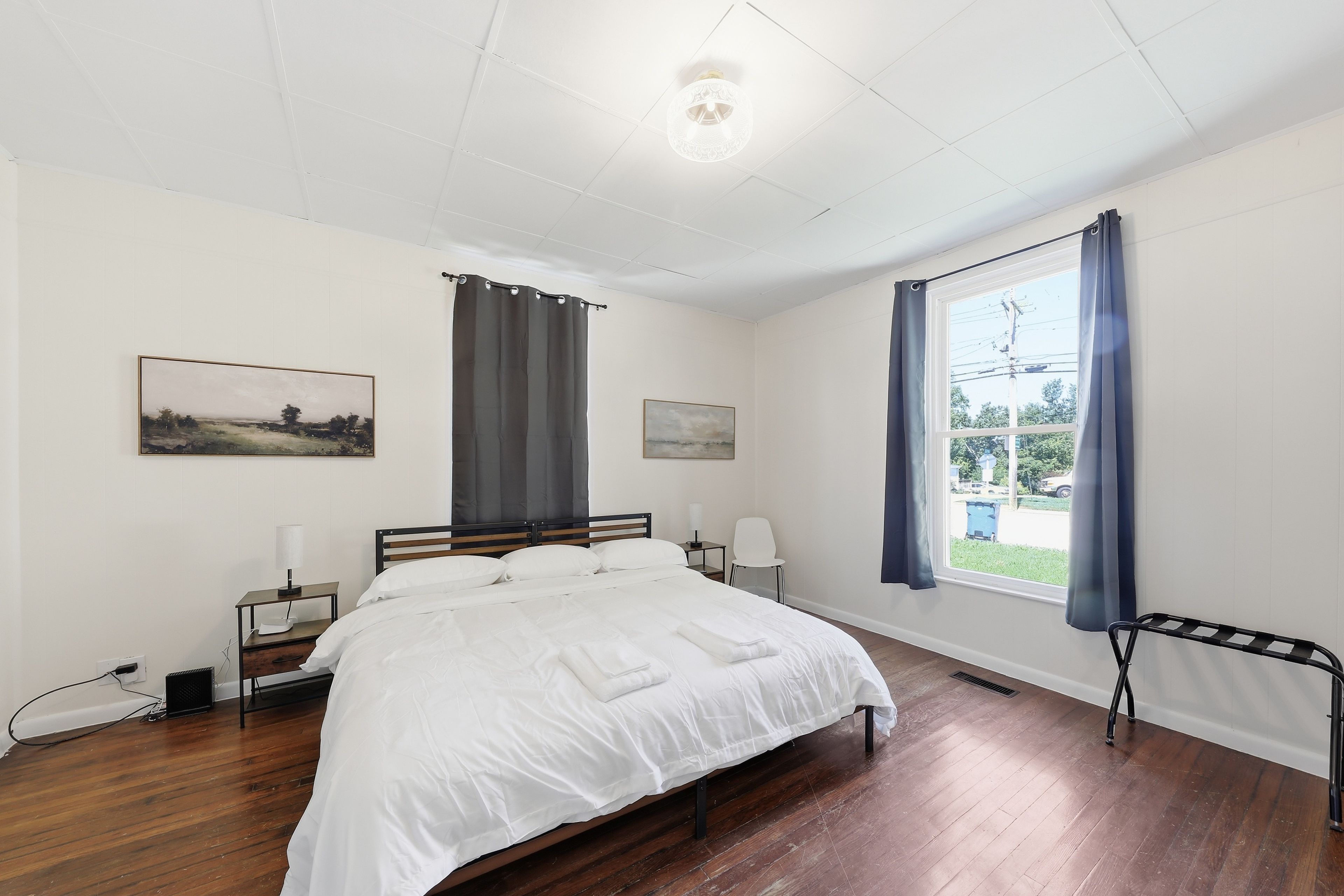 A hotel room with a bed, two bedside tables, a window with curtains, and a painting on the wall.