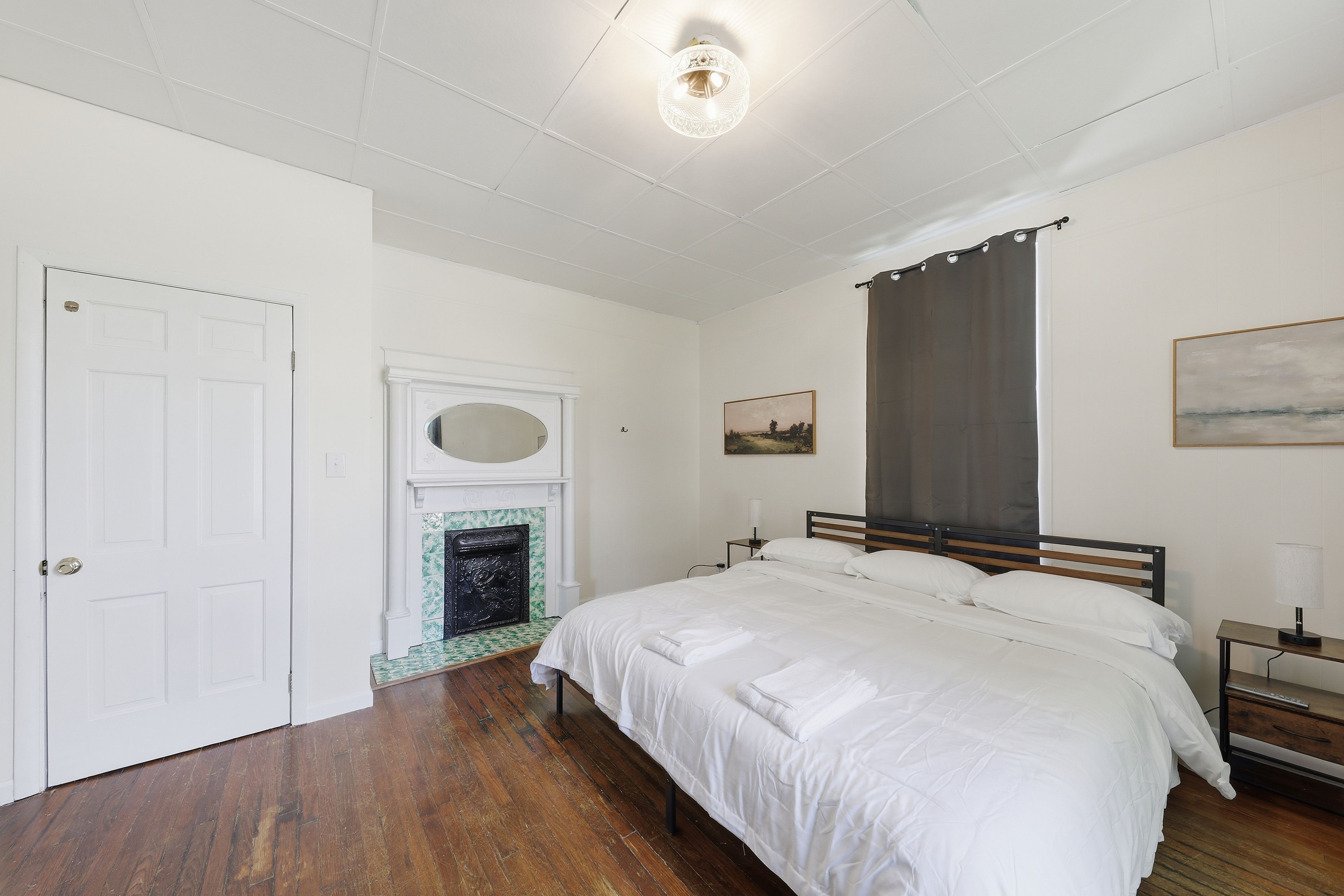 A bedroom with a bed, a nightstand, a lamp, a painting, a fireplace, and a door.