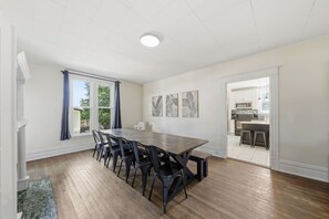 A spacious dining area with a long wooden table and chairs, a kitchen visible through an open doorway, and a ceiling with recessed lighting.