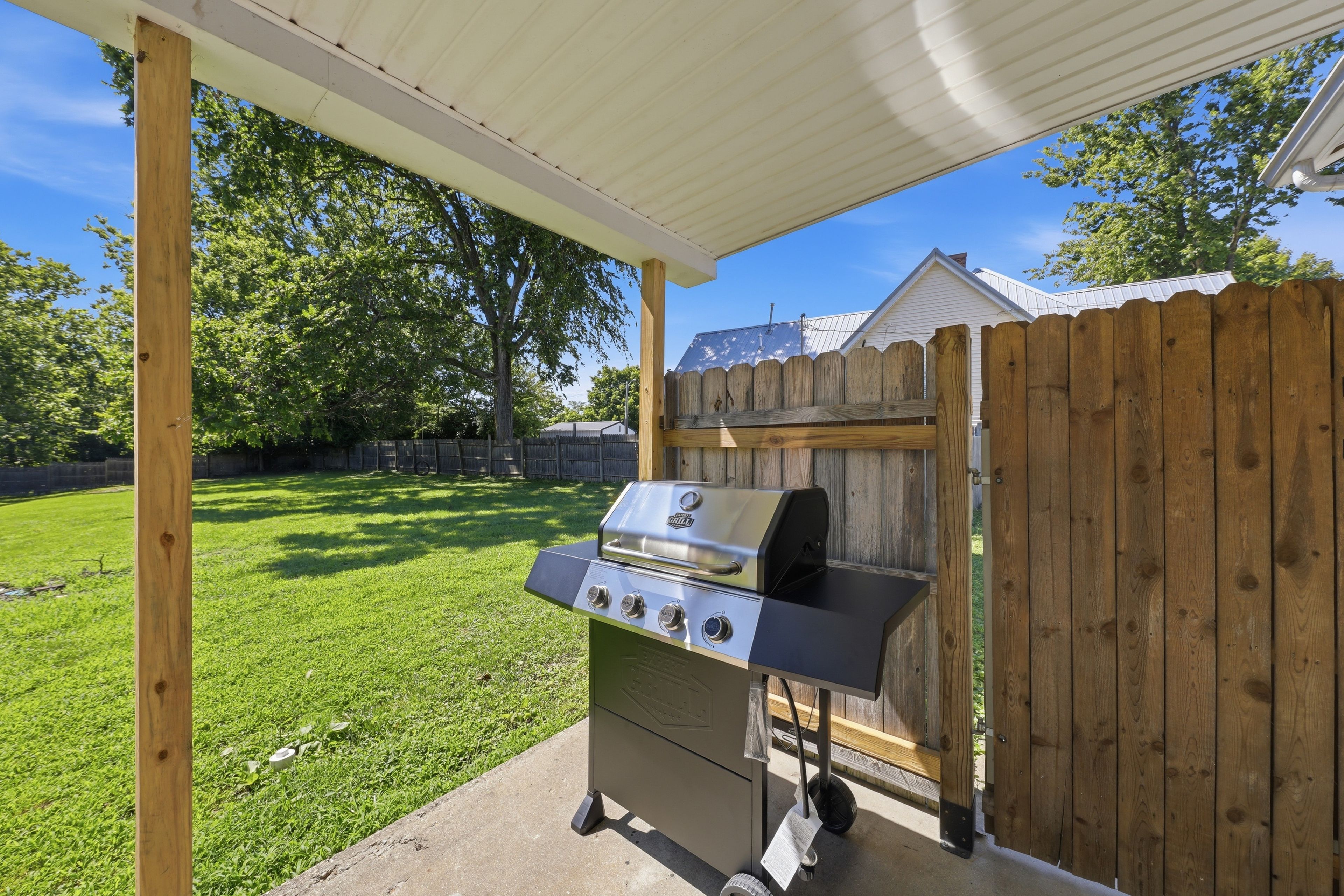 Ein Grill unter einer überdachten Terrasse mit einem Holzzaun im Hintergrund.