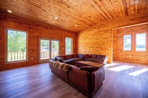 A spacious living room with a large L-shaped sectional sofa, wooden walls, and large windows offering views of the outdoors. - The Chateaugay Lake Mansion (Lyon Mountain)