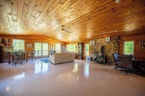 A spacious living area with a wooden ceiling, a stone fireplace, and a large window overlooking greenery. - The Chateaugay Lake Mansion (Lyon Mountain)