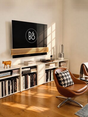 Ein modernes Wohnzimmer mit einem an der Wand befestigten Flachbildfernseher, einem Bücherregal voller Bücher, einem Ledersessel und einer dekorativen Kuhfigur.